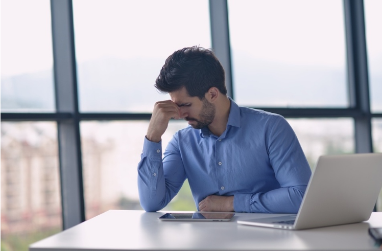 A man in a blue shirt sitting at a desk, looking stressed while holding his head in one hand, with a laptop and a tablet in front of him. The background shows large windows with an outside view.