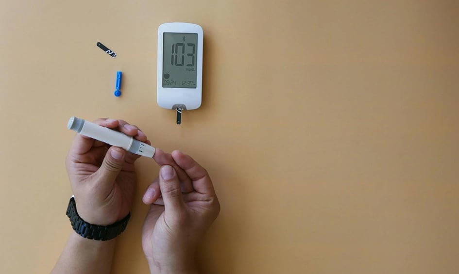 Person using a lancet to test blood glucose levels, with a digital glucometer displaying a reading of 103 mg/dL on a light background.