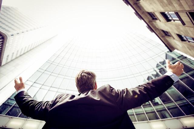 A businessman standing with arms raised toward a towering glass building, symbolizing success or achievement.